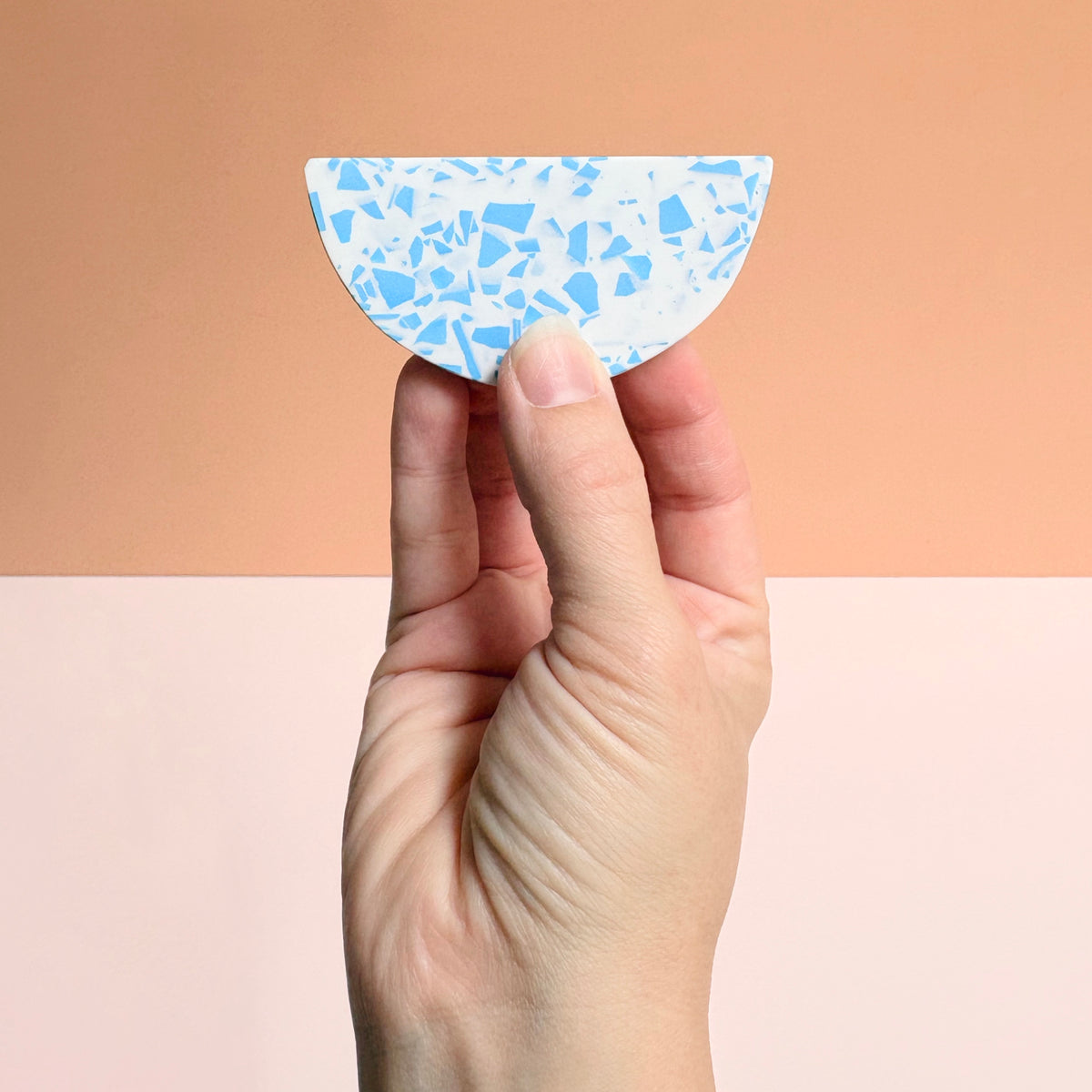 A hand holds a small semi-circular tile with a light blue and white terrazzo pattern against a split peach and pale pink background.