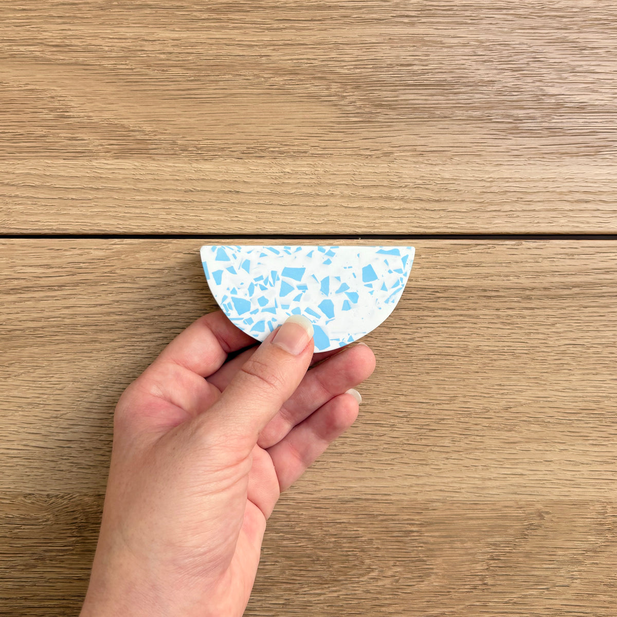 A hand holding a small semicircular drawer pull with a light blue and white terrazzo pattern, set against a light brown wooden background.