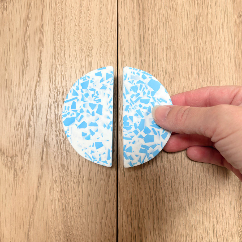A hand holding one half of a round cabinet knob with a blue and white terrazzo pattern, next to the other half resting on a light wood cabinet door.